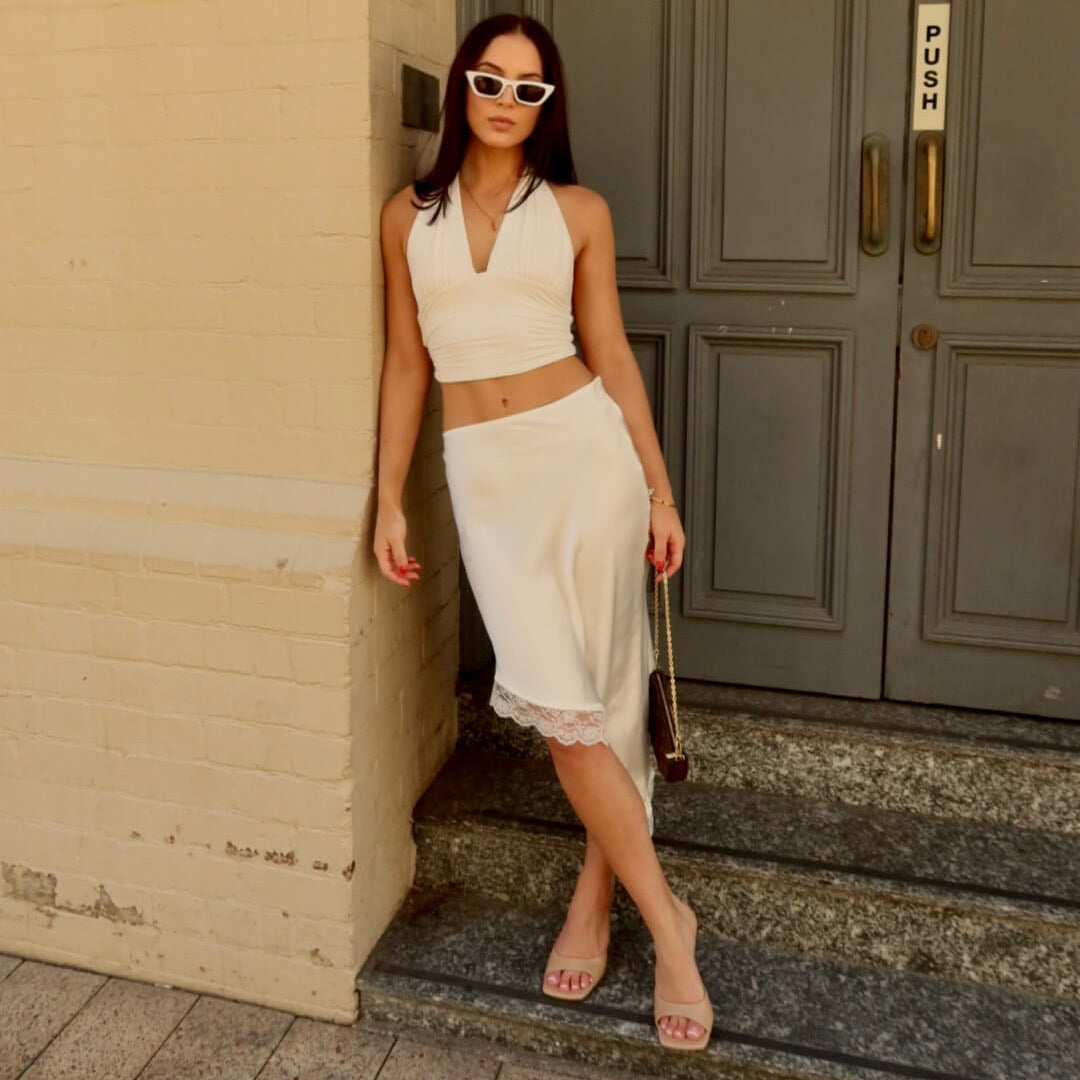 A woman wearing a white sleeveless crop top and matching white midi skirt with lace trim, posing on stone steps in front of a beige brick wall and gray doors.