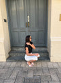 A woman wearing a black short-sleeve top and white pants crouches on the steps in front of a gray double door and cream brick building.
