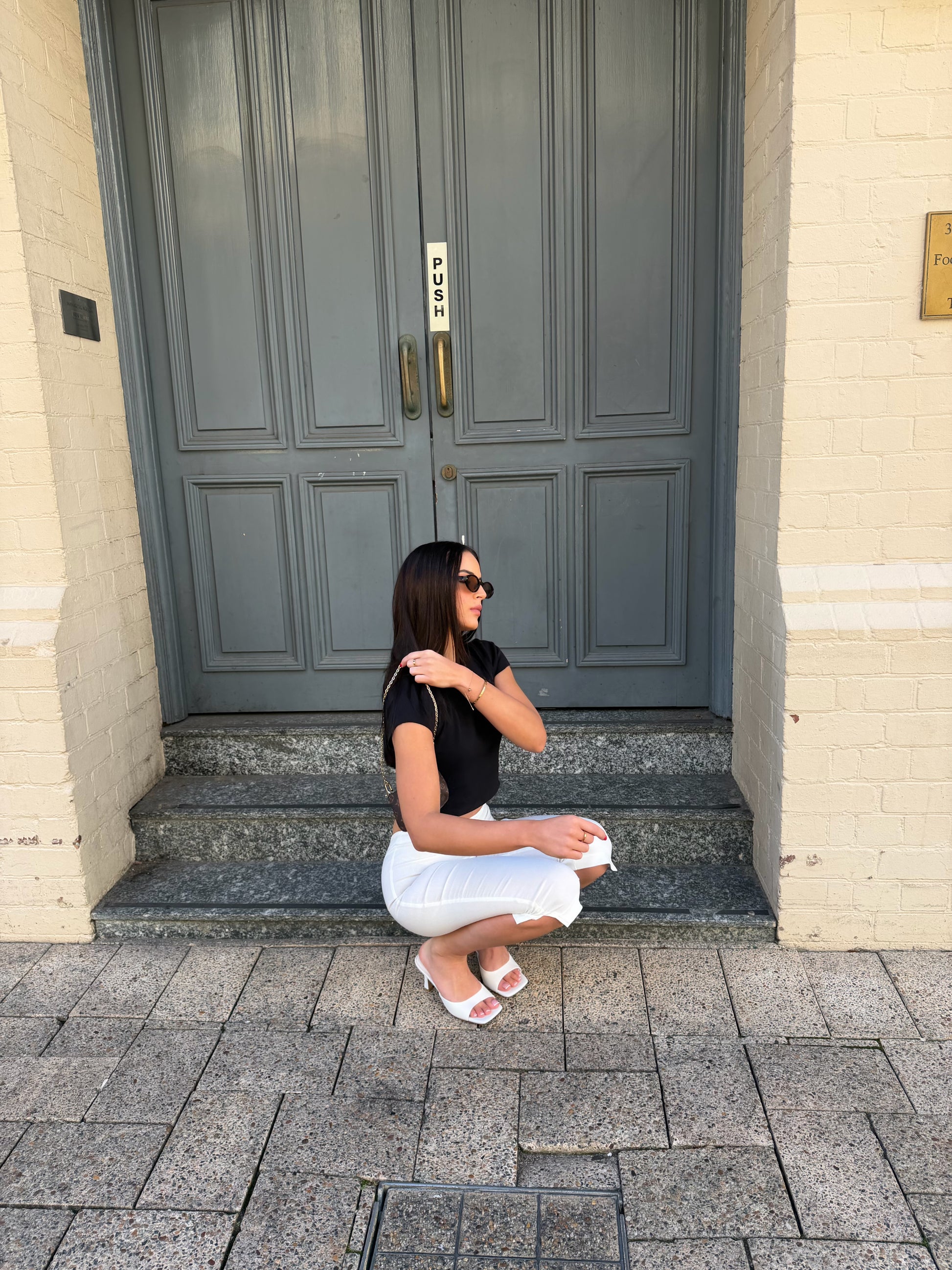 A woman wearing a black short-sleeve top and white pants crouches on the steps in front of a gray double door and cream brick building.